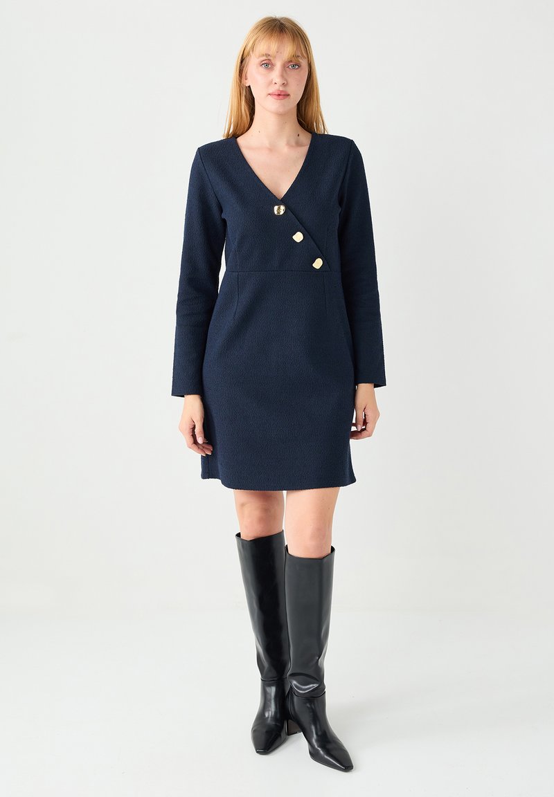 Robe portefeuille bleu marine à manches longues, en tissu texturé, ornée de boutons dorés et d'un décolleté en V. Portée avec des bottes en cuir noir montant jusqu'au genou.