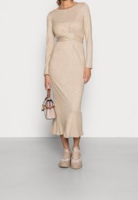 Frau in beigem, langärmligem Midikleid, die eine beige und braune Handtasche hält und beiges Sneakers trägt, steht auf einem grauen Boden.