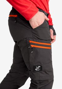 Detaljbild av svarta utomhusbyxor med orange dragkedjefickor och RVRC-logga, bärs av en person i en röd långärmad skjorta med handen i fickan.