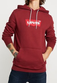 Felpa color bordeaux con tasca a marsupio frontale, lacci bianchi e patch del logo rosso Levi's sul petto. Materiale in cotone, design casual.