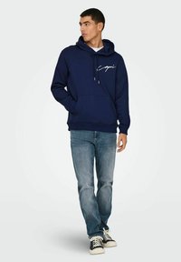 Marineblaue Kapuzenjacke mit frontaler Tasche und Kordeln, ausgestattet mit weißem, geschwungenem Text. Kombiniert mit hellblauen Jeans und dunklen Sneakers.