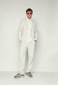 Helle, strukturierte Blazer über einem weißen Shirt, kombiniert mit passender Hose und grauen Sneakers. Sonnenbrillen vervollständigen das Outfit.