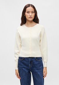 Κρεμ ζακέτα  με στρογγυλή λαιμόκοψη, κούμπωμα με κουμπιά και μακριά μανίκια. Απαλή υφή, εφαρμοστή στη μέση, συνδυασμένη με σκούρα μπλε τζιν.