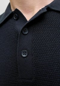 Acercamiento de una camisa de punto negra con textura, tres botones negros en la tapeta del cuello y un cuello doblado.