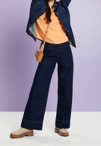 Szerokie, ciemnoniebieskie dżinsy z pomarańczowymi szwami, w połączeniu z miękkim pomarańczowym swetrem i beżowymi botkami na platformie. Mała brązowa torebka na ramię.