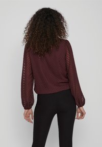 Blouse bordeaux avec un motif en chevrons, manches longues bouffantes et un tissu transparent texturé. Associée à un pantalon noir ajusté.