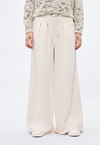 Béžové kalhoty s širokými nohavicemi a stahováním v pase, s plisemi vpředu, měkkou texturou a volným střihem, doplněné o bílé boty.