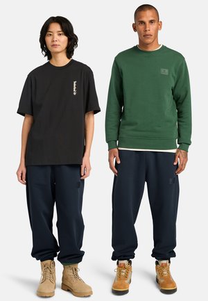 Twee staande modellen met casual outfits: één in een zwarte oversized T-shirt, marineblauwe broek en beige laarzen; de ander in een groene trui, marineblauwe broek en beige wandelschoenen.