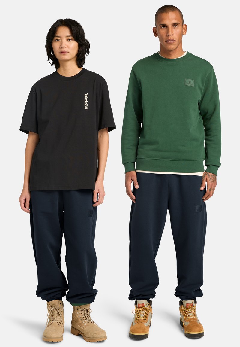 Zwei stehende Models in lässiger Kleidung: Eines trägt ein übergroßes schwarzes T-Shirt, marineblaue Hosen und beige Stiefel; das andere einen grünen Pullover, marineblaue Hosen und beige Wanderschuhe.