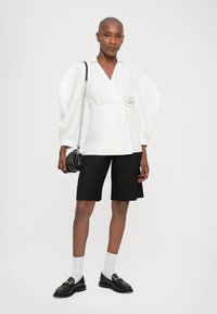 Witte wikkelblouse met lange, volumineuze mouwen, gecombineerd met zwart shorts tot de knieën. Zwarte instappers en witte sokken maken de outfit compleet.