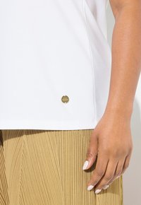 Camiseta blanca de manga corta con una textura suave, que presenta un logo metálico dorado en la parte inferior izquierda. Combinada con pantalones de tela plisada de color beige.