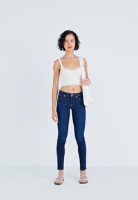 White crop top, dark blue skinny jeans, silver sandals. Model holds a white shoulder bag. Simple background, smooth texture.