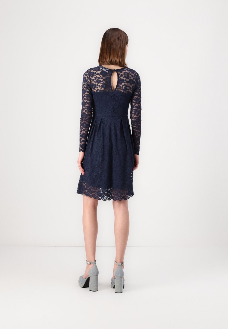 Femme debout, dos à la caméra, portant une robe en dentelle bleu marine à manches longues et des chaussures à talons gris sur fond blanc.