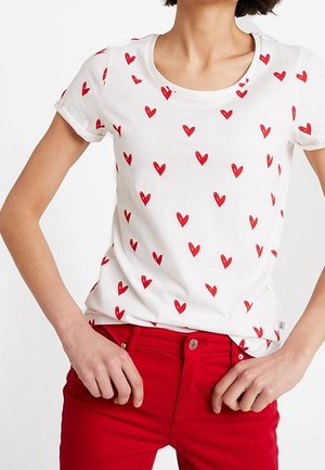 Kobieta ubrana w biały t-shirt z krótkim rękawem z rozrzuconymi czerwonymi sercami i jasnoczerwone spodnie, ręce spoczywają blisko pasa.