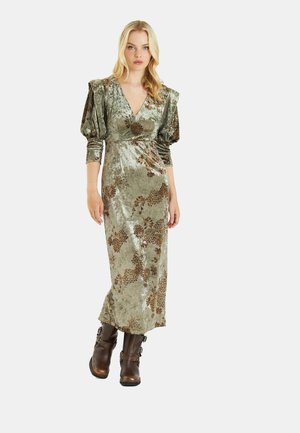 Vestido de terciopelo verde olivo con estampado floral, escote en V profundo, mangas abullonadas y una silueta larga, combinado con botas de tobillo marrones.