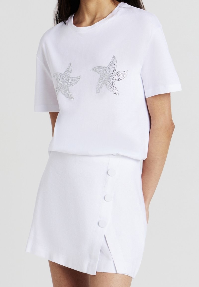 Femme portant une chemise blanche à manches courtes avec des motifs d'étoiles de mer argentées et une jupe blanche avec une fente boutonnée à l'avant.