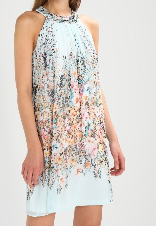 Femme portant une robe sans manches bleu clair avec un motif floral multicolore et un détail plissé à l'avant, debout devant un fond uni.