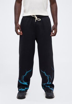 Mann trägt schwarze Jogginghose mit blauen Blitzmustern an den Knöcheln, ein weißes Hemd und schwarze Sneakers, steht vor einem schlichten Hintergrund.