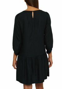 Vestido negro con mangas tres cuartos, cuello redondo y falda con leves pliegues. Presenta un pequeño ojal en la parte posterior con cierre de botón. Textura suave.