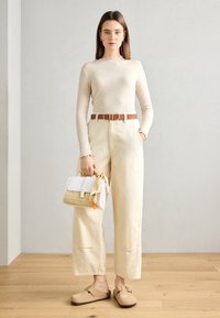 Vrouw staat in een beige geribbelde top met lange mouwen, hoge taille wijde broek, tan riem, instappers, en houdt een witte geweven handtas vast met een zijden sjaal.