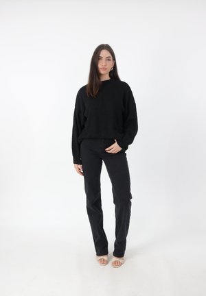 Zwarte oversized gebreide trui met ribbelmanchetten, gecombineerd met hoge taille zwarte jeans. Model staat op een witte achtergrond met open teenschoenen.