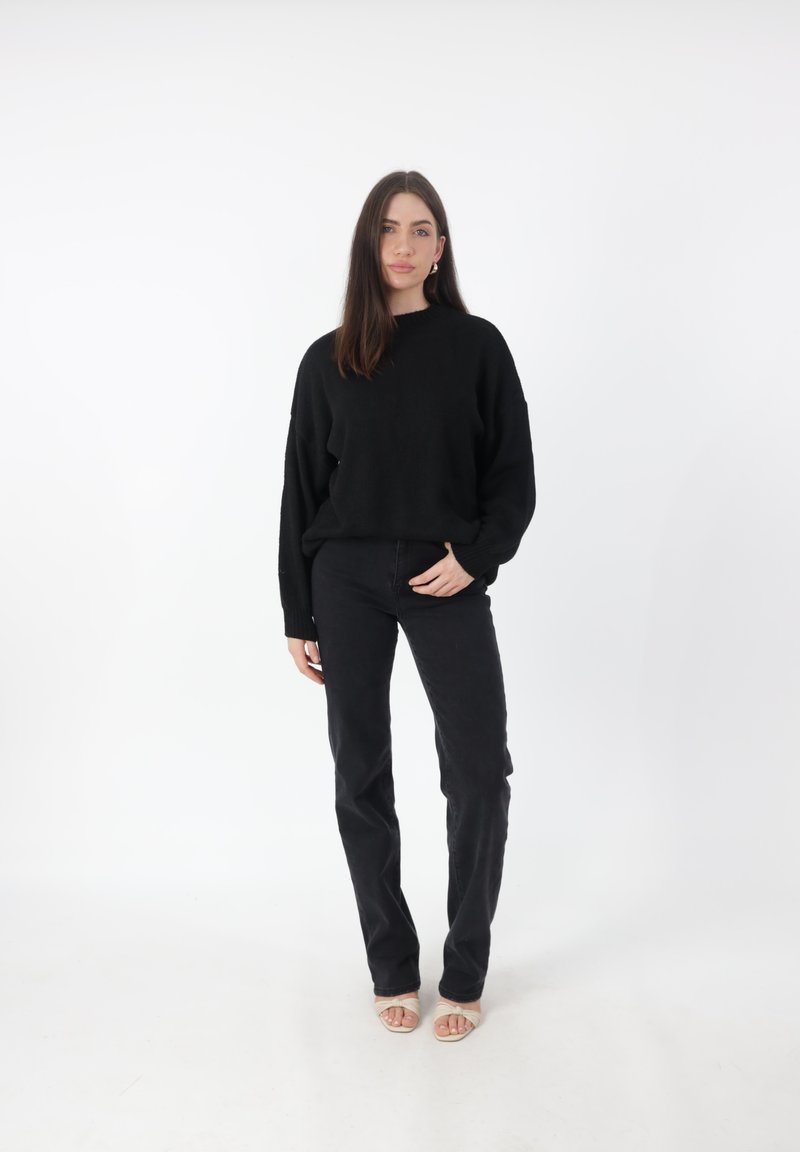 Maglione nero oversize a maglia con polsini a coste, abbinato a jeans neri a vita alta. Il modello si trova su uno sfondo bianco indossando scarpe aperte.