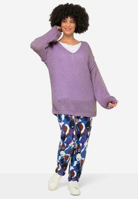 Maglione oversize a collo a V in cotone lavanda, abbinato a pantaloni con motivi geometrici in blu, marrone e nero, con dettagli bianchi.