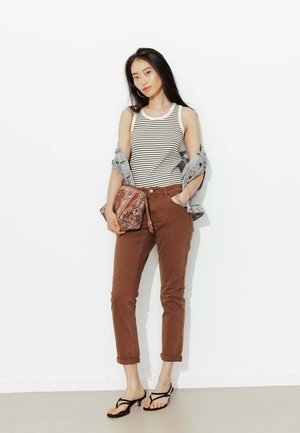 Jeune femme portant un débardeur à rayures, un pantalon marron retroussé, des sandales noires, tenant une pochette marron à motifs, avec une veste en jean posée sur les épaules.