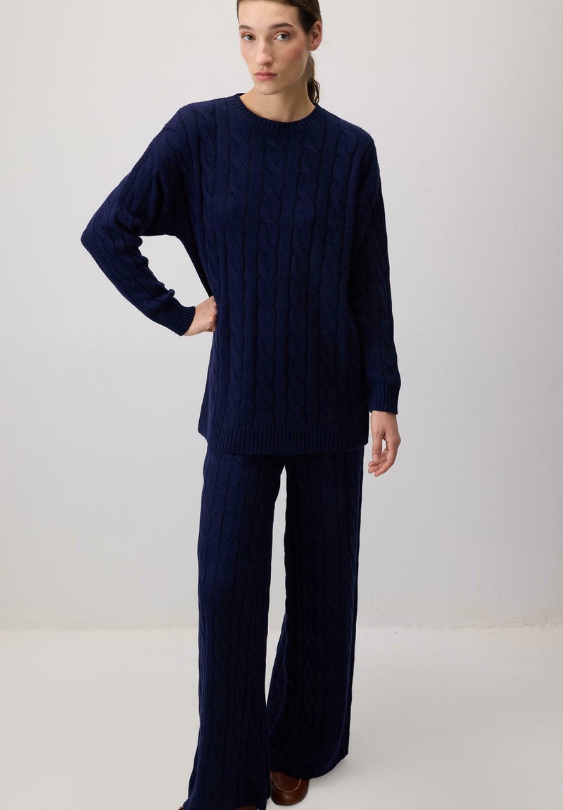 Vrouw staat tegen een effen achtergrond, gekleed in een bijpassende marineblauwe kabelgebreide trui en wijd uitlopende broek met bruine schoenen.