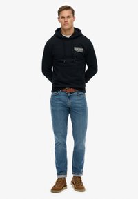 Sudadera con capucha negra de algodón con bolsillo frontal y logo blanco, combinada con vaqueros azules. El conjunto se completa con botas marrones con cordones.