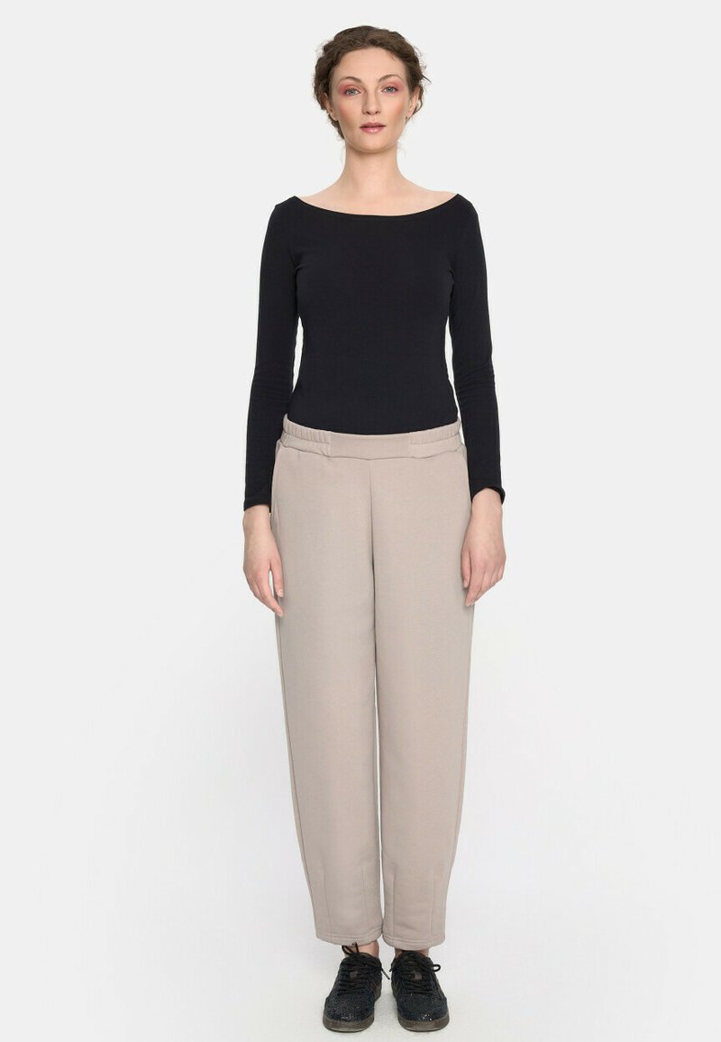 Czarny top z długim rękawem z dekoltem w łódkę, w zestawieniu z beżowymi spodniami o wysokim stanie. Model ma na sobie czarne trampki. Prosty fason, gładka faktura.