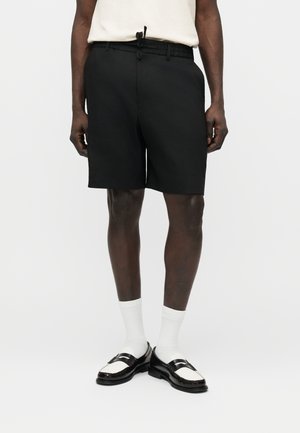 Mann trägt schwarze, maßgeschneiderte Shorts, weiße Socken, schwarz-weiße Loafer und ein weißes Hemd, das in die Hose gesteckt ist, steht vor einem einfarbigen Hintergrund.