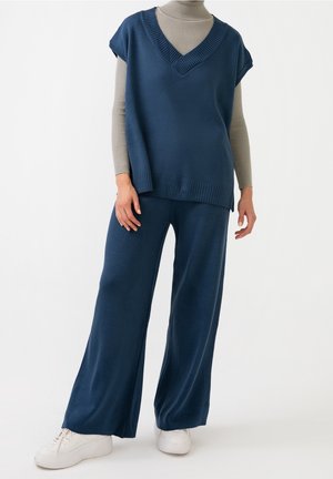Personne portant un pantalon large bleu marine et un pull sans manches à col en V assorti par-dessus un col roulé beige, avec des baskets blanches, debout sur fond blanc.