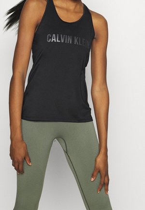 Musta toppi, jossa on kiiltävä printti "CALVIN KLEIN" -logolla. Yhdistetty korkeavyötäröisten oliivinvihreiden legginsien kanssa, joissa on tekstuurisia yksityiskohtia.