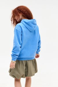 Frau mit lockigem roten Haar, die einen blauen Kapuzenpullover und einen olivgrünen Ballonrock trägt, steht und schaut nach links vor einem weißen Hintergrund.