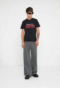Black t-shirt with white and red text reading "Highperformance 1981 REPLAY"; loose gray pants; black chunky shoes; sunglasses.