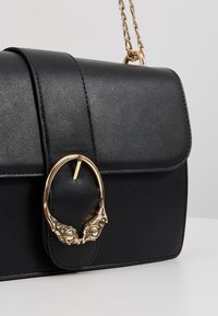 Sac bandoulière en cuir noir avec une bandoulière chaîne dorée et un fermoir décoratif en forme de deux têtes d’animaux qui se touchent.