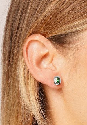 Nahaufnahme eines Ohrs einer Person mit glattem hellbraunem Haar, die einen silbernen Steckerohrring mit einem rechteckigen grünen Edelstein trägt.