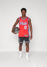 Maglia da basket rossa con accenti blu e testo "Phila". Pantaloni corti neri con strisce bianche. Tiene un pallone da basket nero. Scarpe sportive bianche.