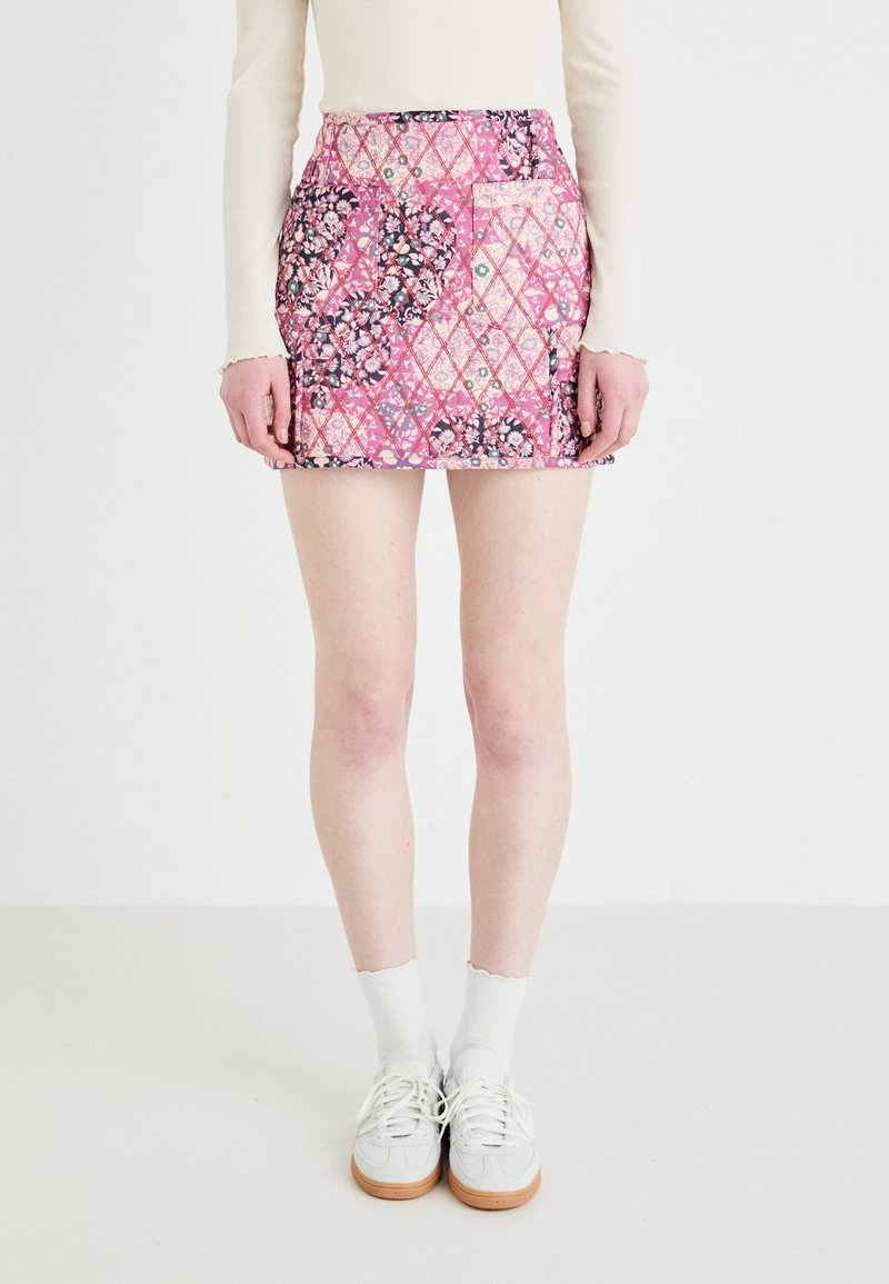 Personne portant une mini-jupe fleurie rose avec un design patchwork, des chaussettes blanches, des baskets blanches et un haut à manches longues crème sur un fond uni.