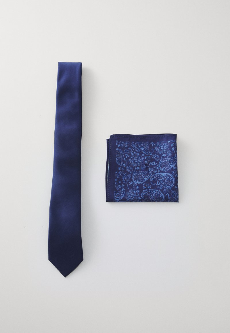 Tengerészsötét selyemnyakkendő egy illő zsebkendő mellett, amely különböző kék árnyalatú paisley mintát mutat. Mindkét darab gondosan elrendezve egy fehér háttéren.