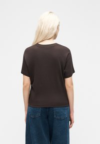 Ξανθιά γυναίκα με την πλάτη στραμμένη, φοράει ένα απλό σκούρο καφέ κοντομάνικο μπλουζάκι και φαρδιά μπλε τζιν, μπροστά σε ανοιχτό φόντο.