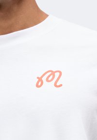 Κοντινή λήψη ενός λευκού T-shirt με ένα μικρό πορτοκαλί ζώδιο Λέοντα τυπωμένο στο στήθος κοντά στη λαιμόκοψη.