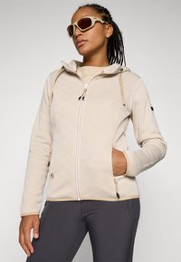 Hoodie a motivo color beige chiaro con zip, cappuccio regolabile e tasche laterali. Indossato con pantaloni scuri e abbinato a occhiali da sole chiari.