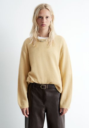 Gelber Strickpullover mit langen Ärmeln und rundem Halsauschnitt, kombiniert mit braunen Lederhosen und einer schlichten Metallkette.