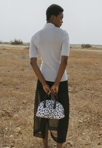 Maža rankinė juodos ir baltos spalvų, su dėmelių raštu, laikoma žmogaus, apsirengusio šviesiai pilku marškinėliu ir ilga juoda sijonu.