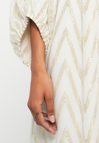 Tecido branco com padrão zig-zag em dourado metálico; apresenta mangas bufantes com detalhe franzido. Mão posicionada perto da superfície texturizada.