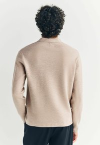 Beige stickad tröja med långa ärmar och krage. Har en texturerad finish och en rak fåll, bärs över mörka byxor.