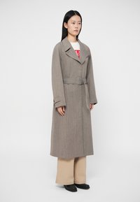 Γκρι παλτό με ζώνη, φαρδιά γιακά και κάθετο ριγέ μοτίβο, συνδυασμένο με μπεζ φαρδιά παντελόνια και μαύρα παπούτσια.