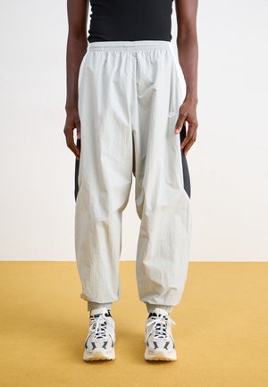 Persoon draagt losse lichtgrijze Nike trainingsbroek, witte en zwarte sneakers en een zwart mouwloos topje, staand op een gele vloer.